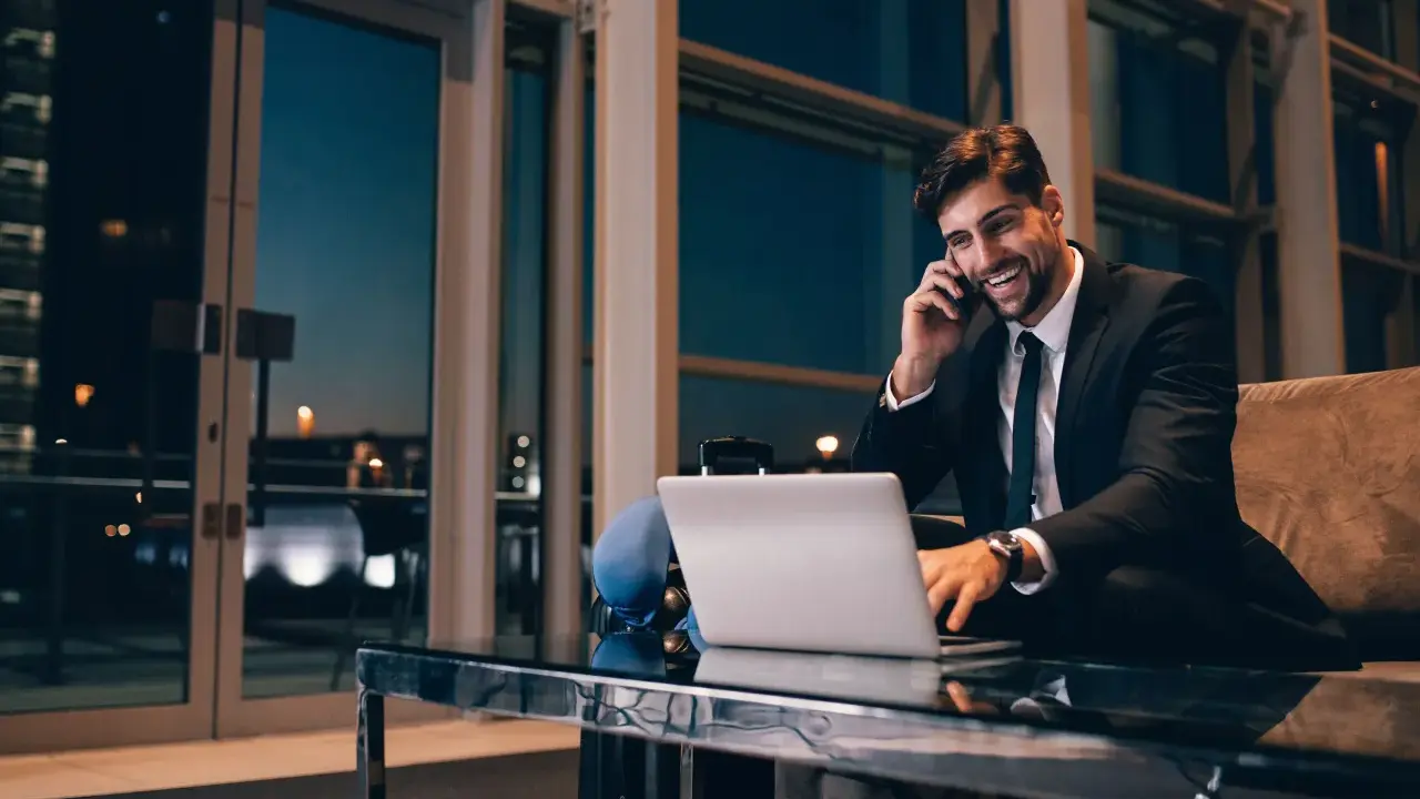 Uśmiechnięty mężczyzna w garniturze rozmawia przez telefon, pracując przy laptopie. Czas na rozliczenie delegacji.