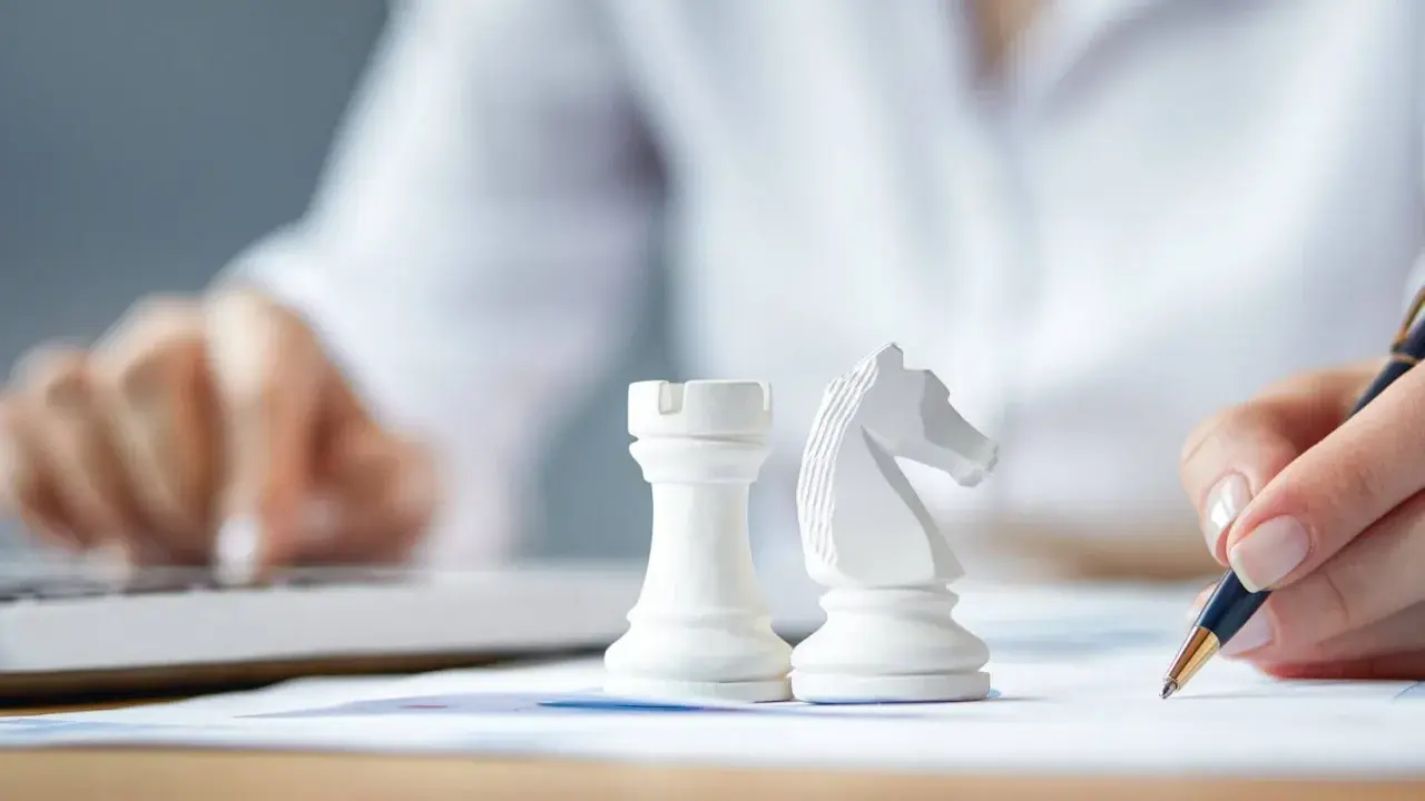 Strategia dla dużych firm: białe figury szachowe, ręka pisząca na papierze, druga ręka na klawiaturze laptopa.