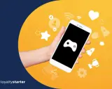 Dłoń trzyma smartfon z ikoną gamepada. Wokół ikony symbolizujące gamifikację przykłady: gwiazdki, serce, rakiety, trofeum.