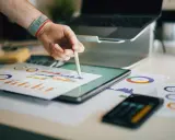 Ręka z rysikiem analizuje wykresy na biurku, gdzie znajduje się tablet, laptop i dokumenty. To idealne miejsce do pracy nad strategią e commerce magazyn.