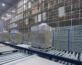 Przenośniki magazynowe transportują palety z towarami w nowoczesnym magazynie. Wysokie regały wypełnione są produktami.