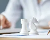Strategia dla dużych firm: białe figury szachowe, ręka pisząca na papierze, druga ręka na klawiaturze laptopa.