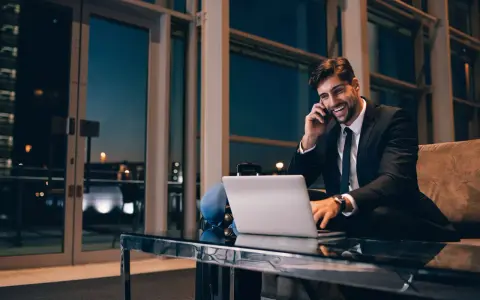 Uśmiechnięty mężczyzna w garniturze rozmawia przez telefon, pracując przy laptopie. Czas na rozliczenie delegacji.