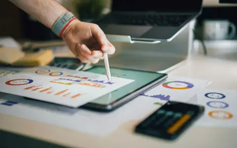 Ręka z rysikiem analizuje wykresy na biurku, gdzie znajduje się tablet, laptop i dokumenty. To idealne miejsce do pracy nad strategią e commerce magazyn.