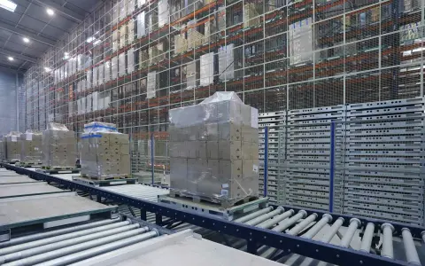 Przenośniki magazynowe transportują palety z towarami w nowoczesnym magazynie. Wysokie regały wypełnione są produktami.