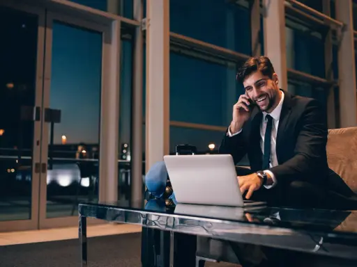 Uśmiechnięty mężczyzna w garniturze rozmawia przez telefon, pracując przy laptopie. Czas na rozliczenie delegacji.