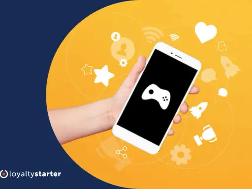 Dłoń trzyma smartfon z ikoną gamepada. Wokół ikony symbolizujące gamifikację przykłady: gwiazdki, serce, rakiety, trofeum.