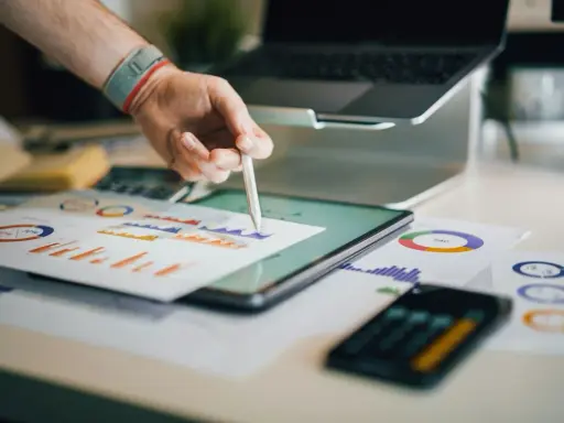 Ręka z rysikiem analizuje wykresy na biurku, gdzie znajduje się tablet, laptop i dokumenty. To idealne miejsce do pracy nad strategią e commerce magazyn.