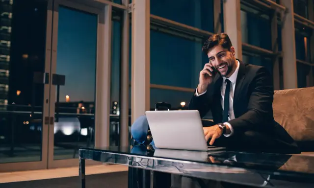 Uśmiechnięty mężczyzna w garniturze rozmawia przez telefon, pracując przy laptopie. Czas na rozliczenie delegacji.