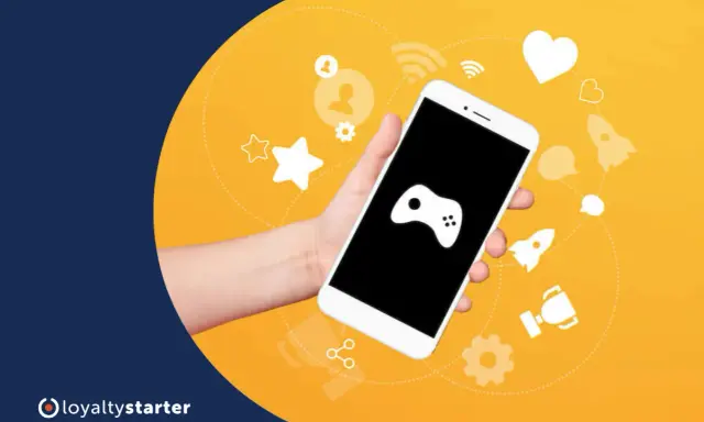 Dłoń trzyma smartfon z ikoną gamepada. Wokół ikony symbolizujące gamifikację przykłady: gwiazdki, serce, rakiety, trofeum.