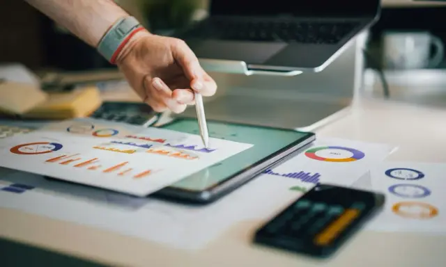 Ręka z rysikiem analizuje wykresy na biurku, gdzie znajduje się tablet, laptop i dokumenty. To idealne miejsce do pracy nad strategią e commerce magazyn.