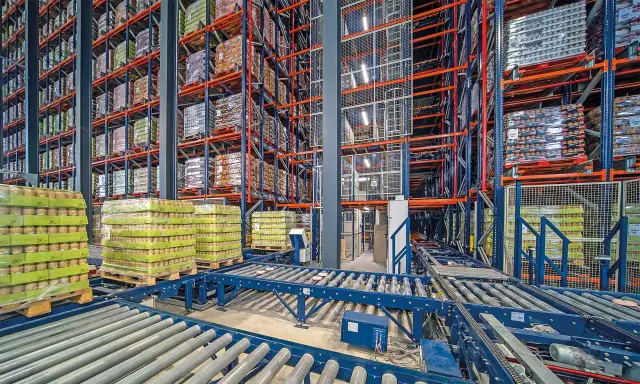 Wysoki magazyn z regałami automatycznymi, pełen towarów na paletach. Taśmy transportowe przenoszą ładunki.