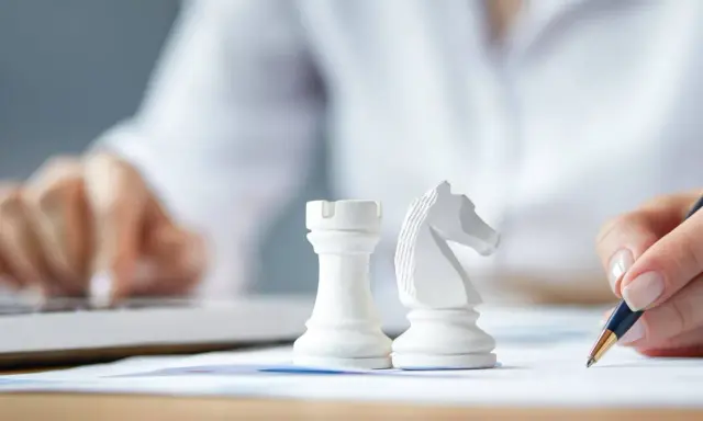 Strategia dla dużych firm: białe figury szachowe, ręka pisząca na papierze, druga ręka na klawiaturze laptopa.