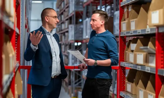 Dwóch mężczyzn dyskutuje w dziale magazynowym, otoczeni regałami z paczkami. Jeden z nich gestykuluje, drugi trzyma dokumenty.