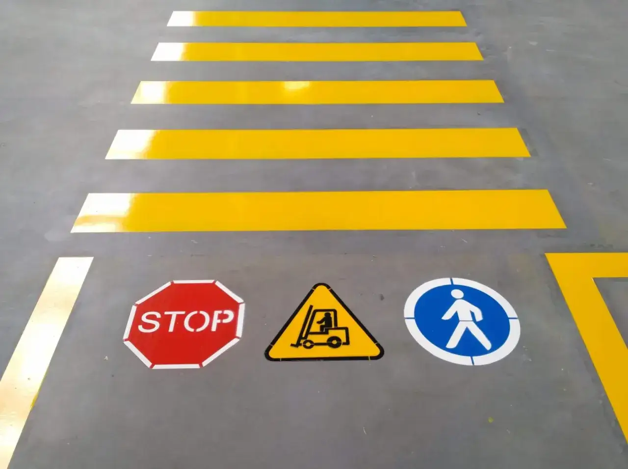 Oznakowane drogi transportowe w magazynie: pasy dla pieszych, znak STOP, ostrzeżenie o wózku widłowym i ścieżka dla pieszych.